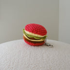 A back view of a small crocheted hamburger keychain with a red top bun, green lettuce, and brown patty, resting on a textured white surface against a plain light gray background.