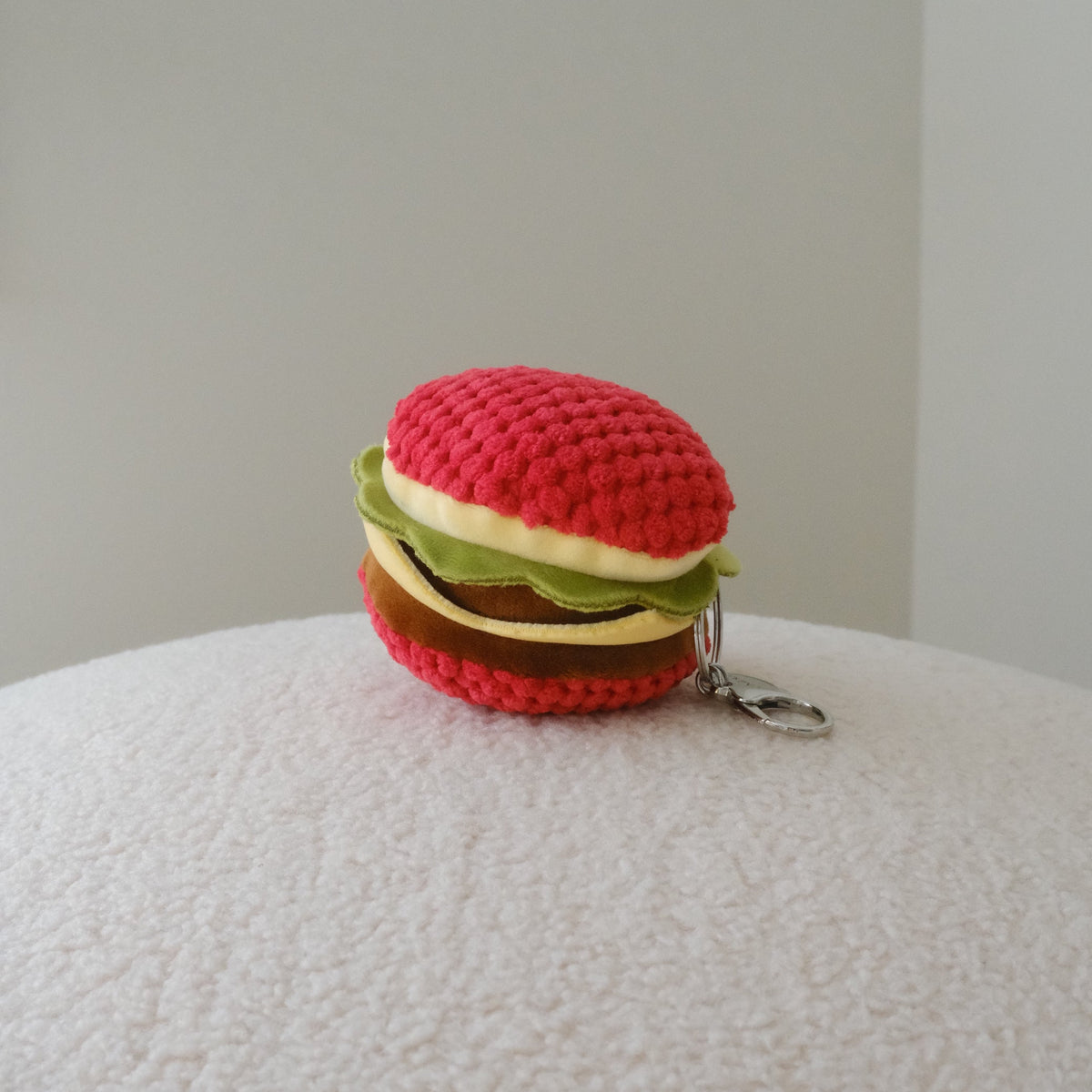 A back view of a small crocheted hamburger keychain with a red top bun, green lettuce, and brown patty, resting on a textured white surface against a plain light gray background.