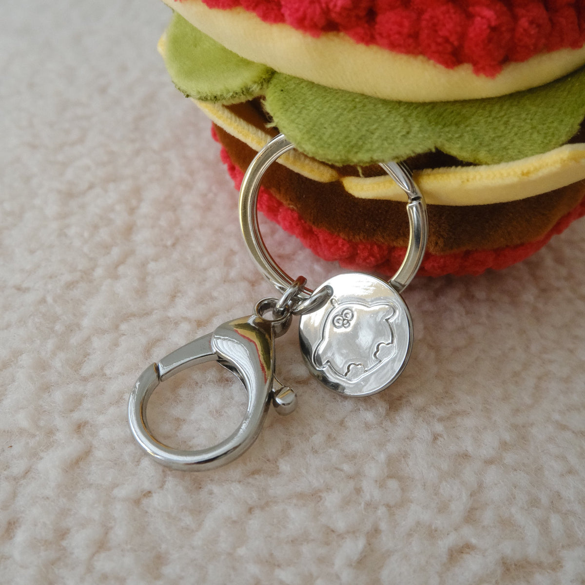 A close-up of a plush toy shaped like a hamburger with a silver metal keychain attached. The toy features a red textured top, yellow sides, green middle layer, and brown bottom layer, resting on a soft beige textured surface.