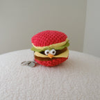 A small crocheted plush keychain shaped like a hamburger with a cute face, resting on a textured white surface against a neutral light gray background.
