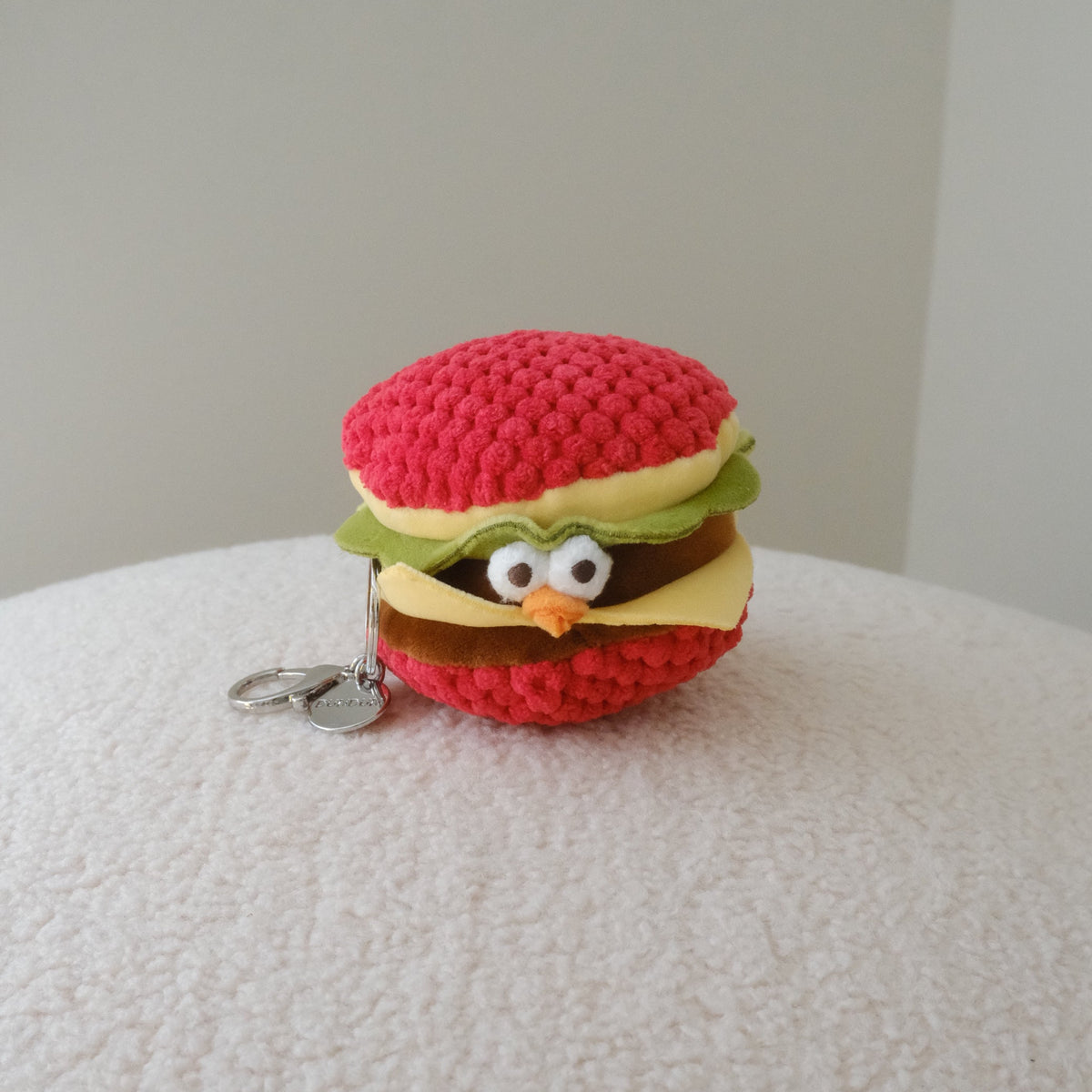 A small crocheted plush keychain shaped like a hamburger with a cute face, resting on a textured white surface against a neutral light gray background.