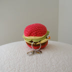 A small crocheted keychain shaped like a hamburger, with a red top bun, green lettuce, yellow cheese, brown bun, and silver metal keyring, resting on a textured white surface against a plain light gray background.
