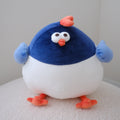 A round, puffy plush toy bird with a dark blue top half, white belly, light blue wings, and orange feet and beak, sitting on a white textured surface against a plain light gray wall.