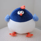 A round, puffy plush toy bird with a dark blue top half, white belly, light blue wings, and orange feet and beak, sitting on a white textured surface against a plain light gray wall.