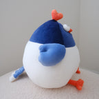 A plush toy bird with a white body, blue wing and head, and orange feet and beak, sitting on a textured cream surface against a plain light gray wall.