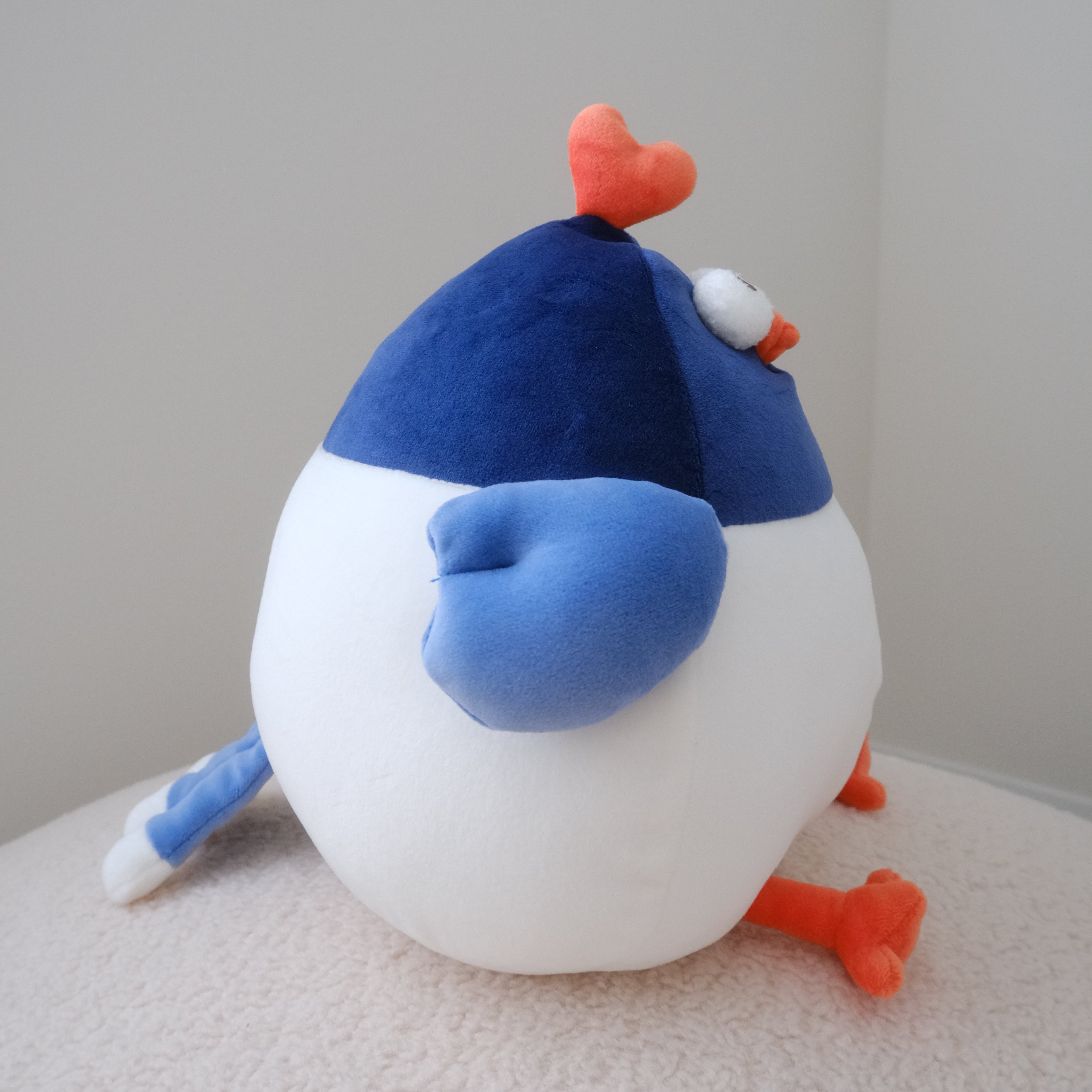 A plush toy bird with a white body, blue wing and head, and orange feet and beak, sitting on a textured cream surface against a plain light gray wall.