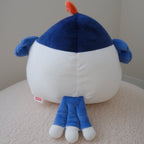 A blue and white plush toy with a small orange chicken crown on top and blue wings photographed from the back while sitting on a beige textured surface against a plain light gray wall.