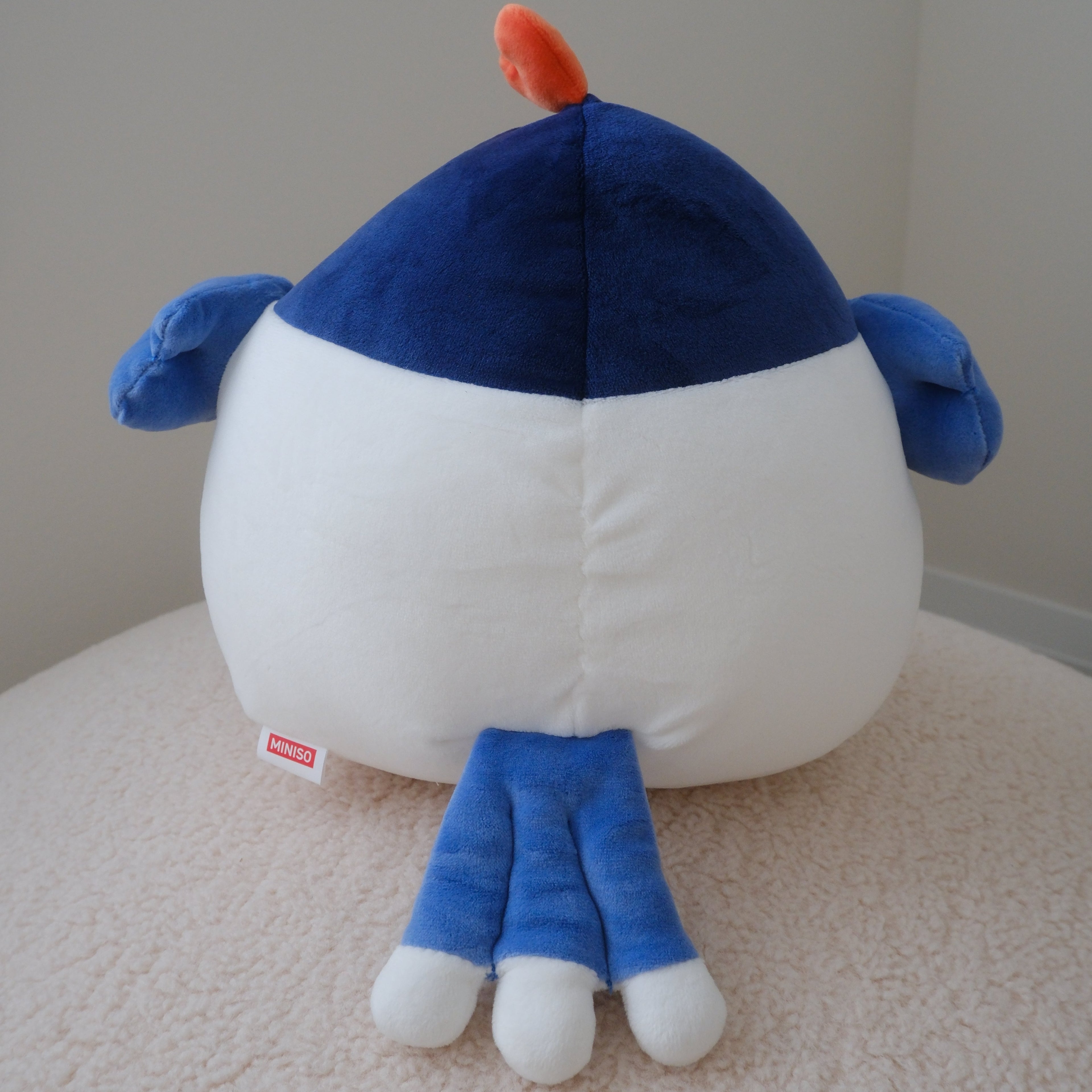 A blue and white plush toy with a small orange chicken crown on top and blue wings photographed from the back while sitting on a beige textured surface against a plain light gray wall.