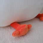 Close-up of a plush toy with a large white body and bright orange feet resting on a soft cream-colored textured surface.