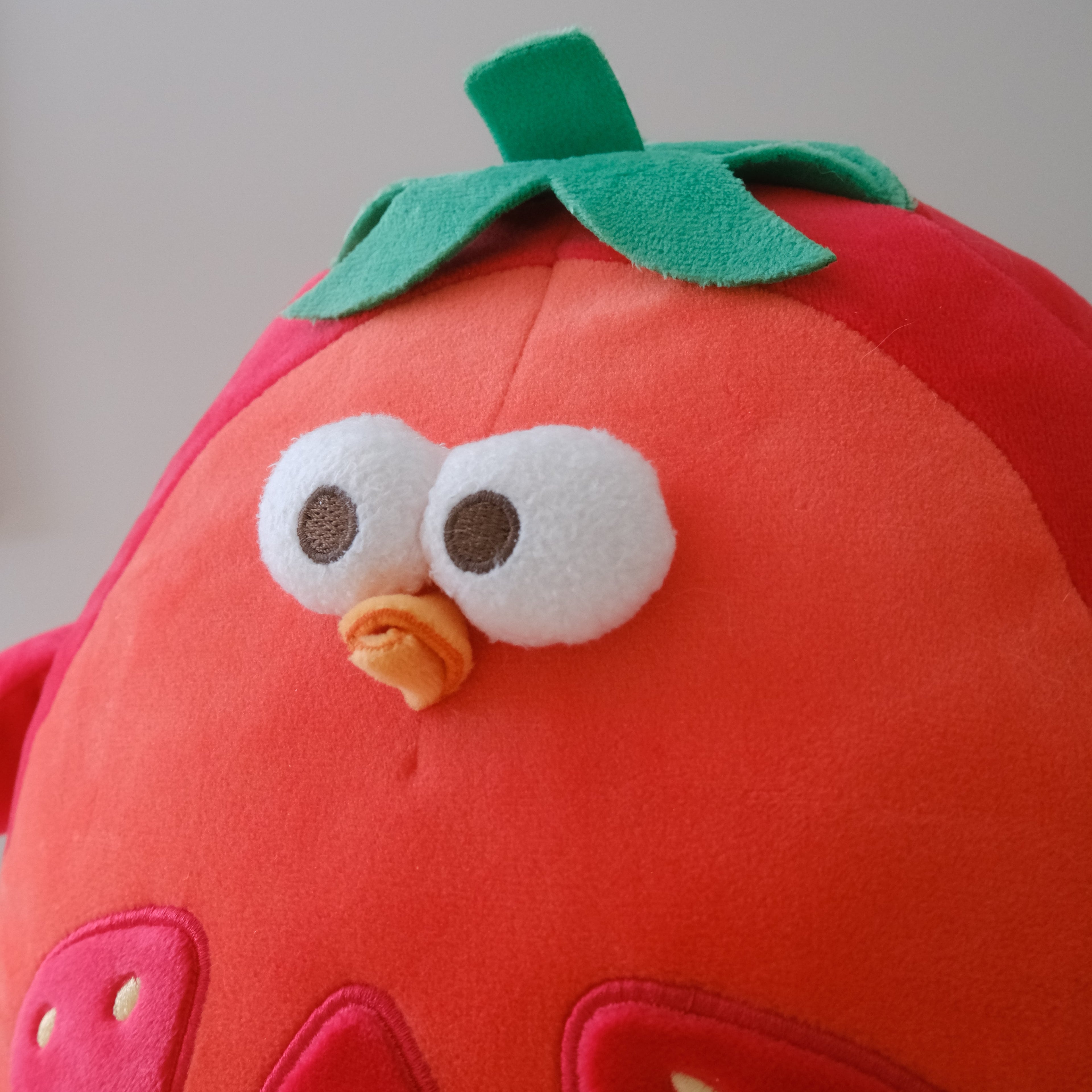 A close-up of a red plush tomato toy with a green felt leafy top, large white embroidered eyes, and a small orange stitched beak.