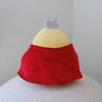 A plush chicken toy with a yellow body and red cape, displayed against a plain light gray wall on a white textured surface.