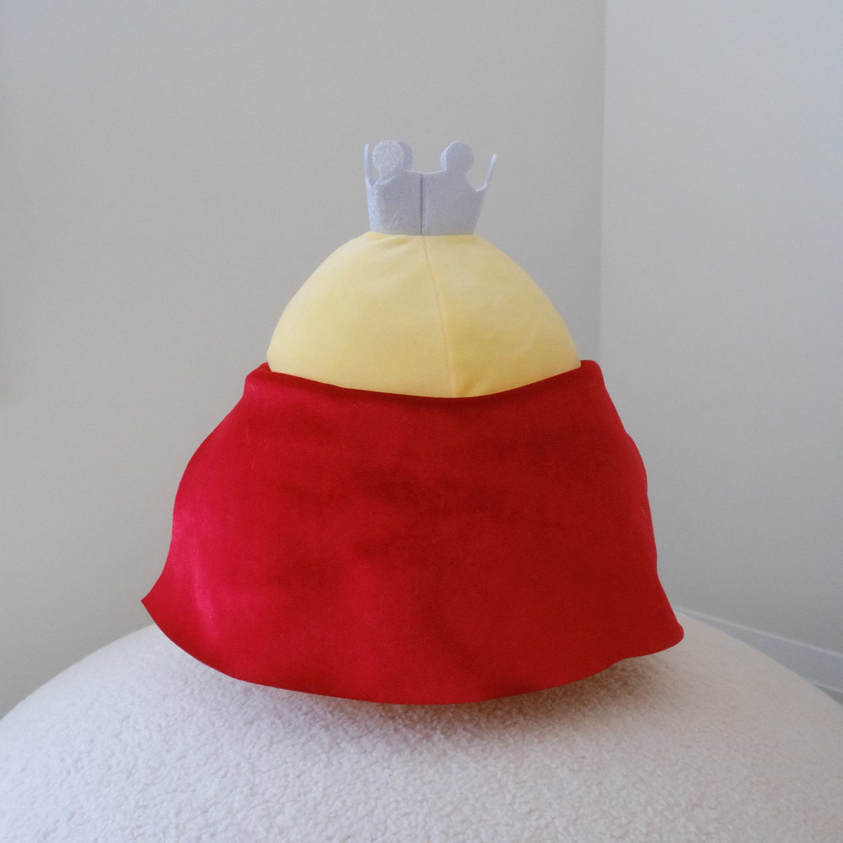 A plush chicken toy with a yellow body and red cape, displayed against a plain light gray wall on a white textured surface.