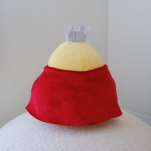 A plush chicken toy with a yellow body and red cape, displayed against a plain light gray wall on a white textured surface.