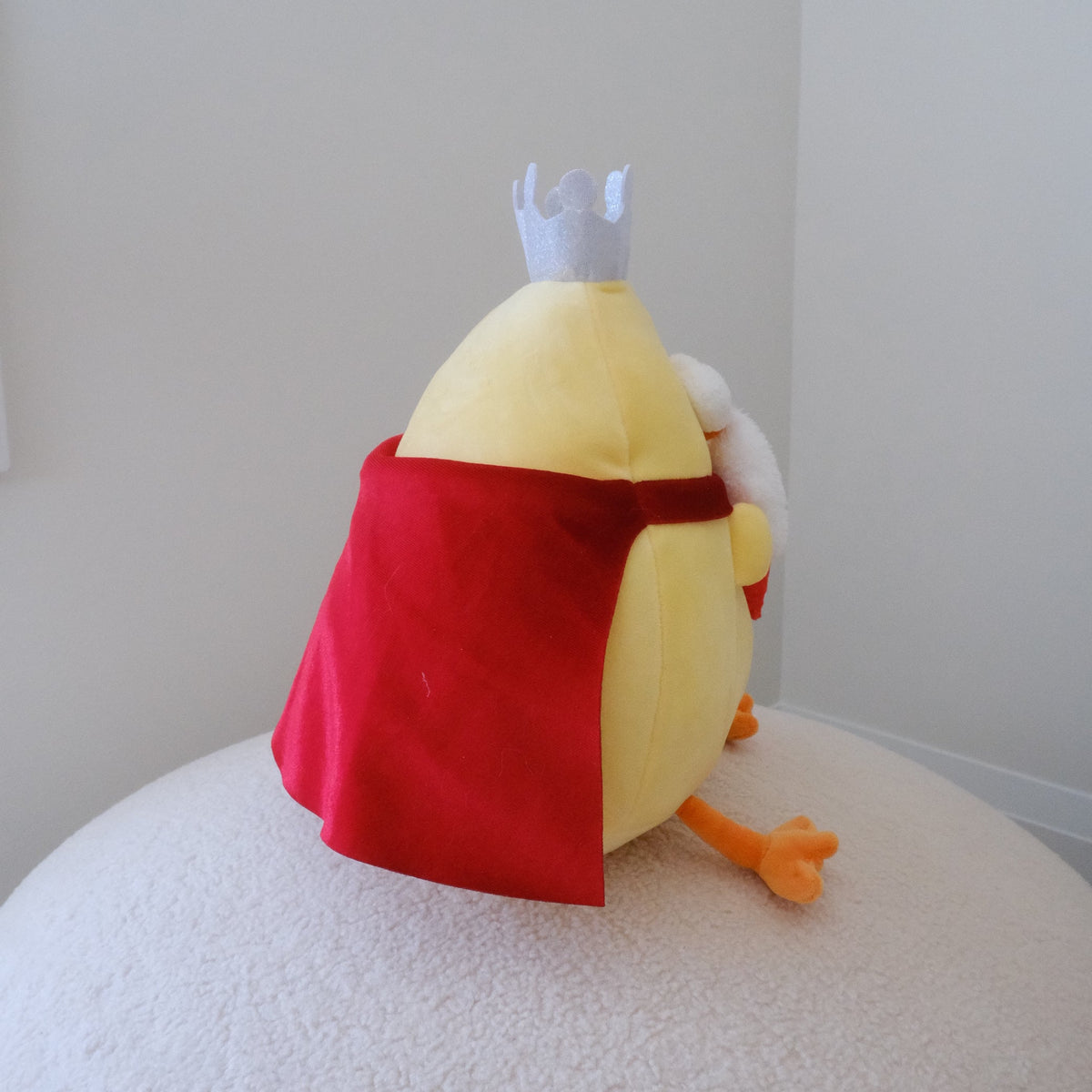 A plush toy chicken dressed as a king, featuring a yellow body, red cape, white crown, and orange beak/legs, sitting on a textured white surface against a plain light gray wall.