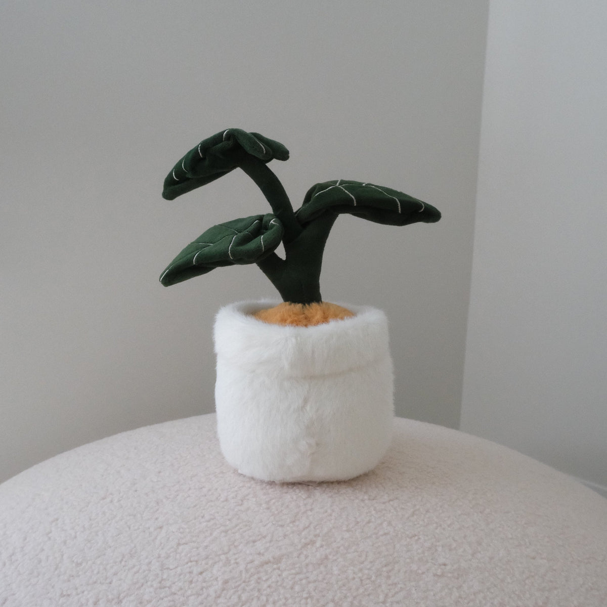 A small plush plant toy with dark green leaves and a white fuzzy pot, placed on a round textured surface against a plain light gray wall.
