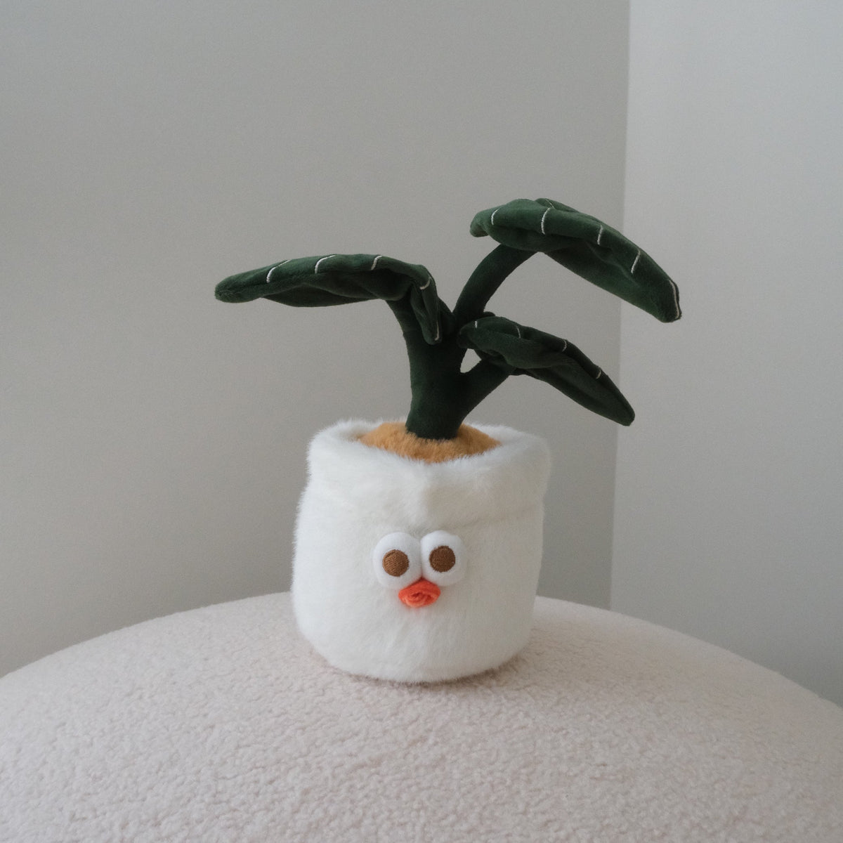 A small plush plant toy with dark green leaves and a white fuzzy pot featuring a cute face, displayed on a textured cream surface against a neutral light gray background.
