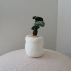A small plush monstera toy in a fuzzy white pot, displayed on a textured cream surface against a plain light gray wall.