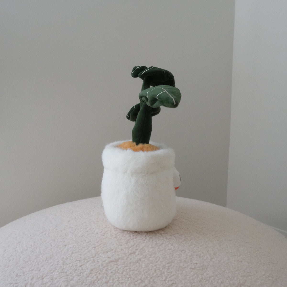 A small plush monstera toy in a fuzzy white pot, displayed on a textured cream surface against a plain light gray wall.