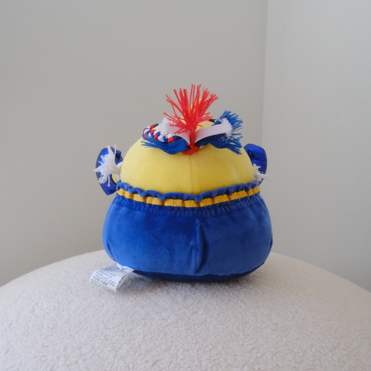 A plush toy shaped like a yellow chicken, featuring blue pants, and a red and white braided headband, sitting on a textured white surface against a plain light gray wall.