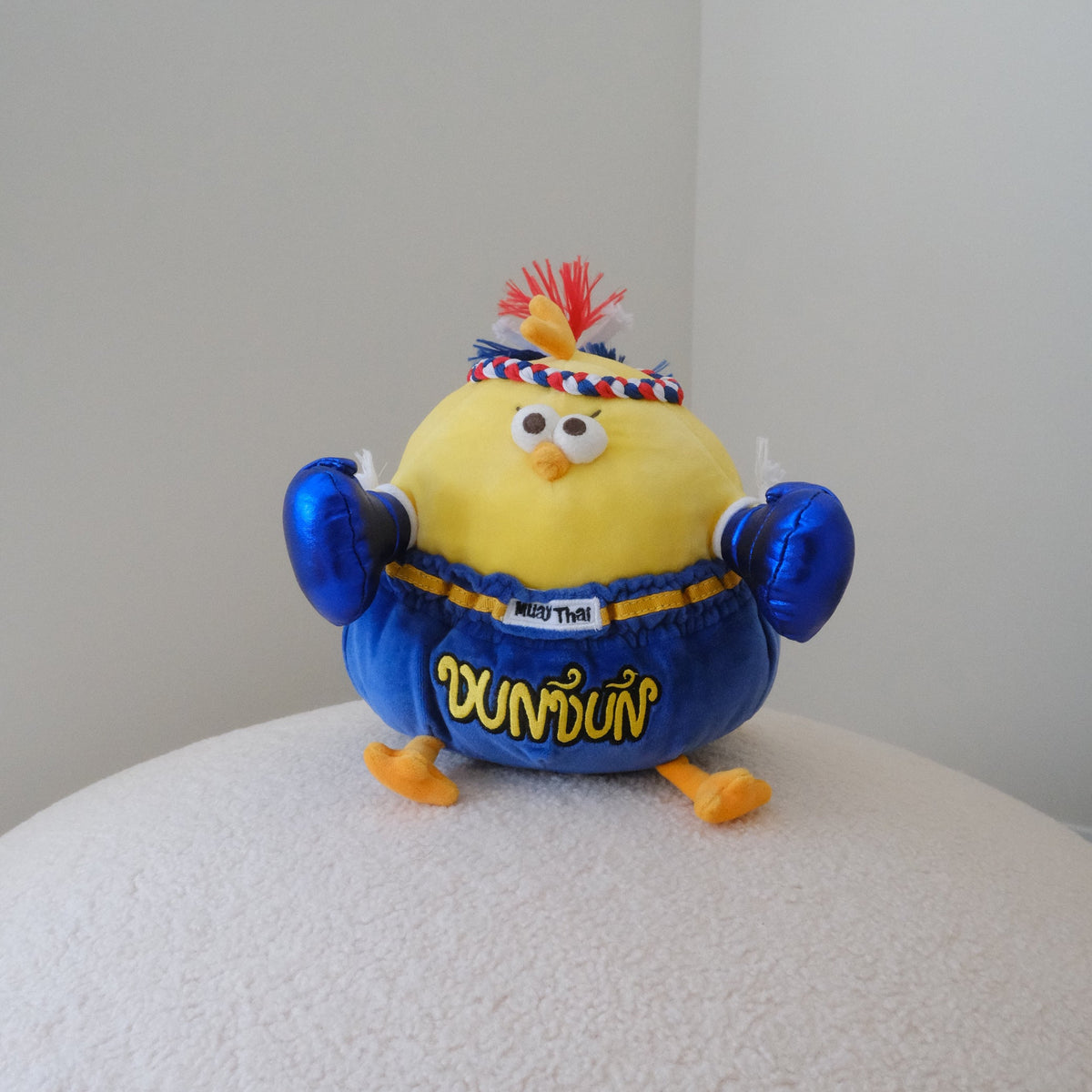 A yellow plush toy chick wearing a blue muay thai outfit with 'DUNDUN' written on the front, standing on a textured white surface against a plain light gray background.
