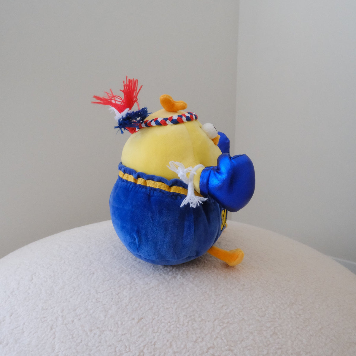 A small plush toy shaped like a yellow chick wearing blue pants and blue boxing gloves, sitting on a textured white surface against a plain light gray wall.