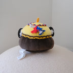 A plush toy shaped like a chicken, featuring a dark brown shorts, a yellow body, and a red, white, and blue braided headband, displayed on a textured cream surface against a plain light gray background.