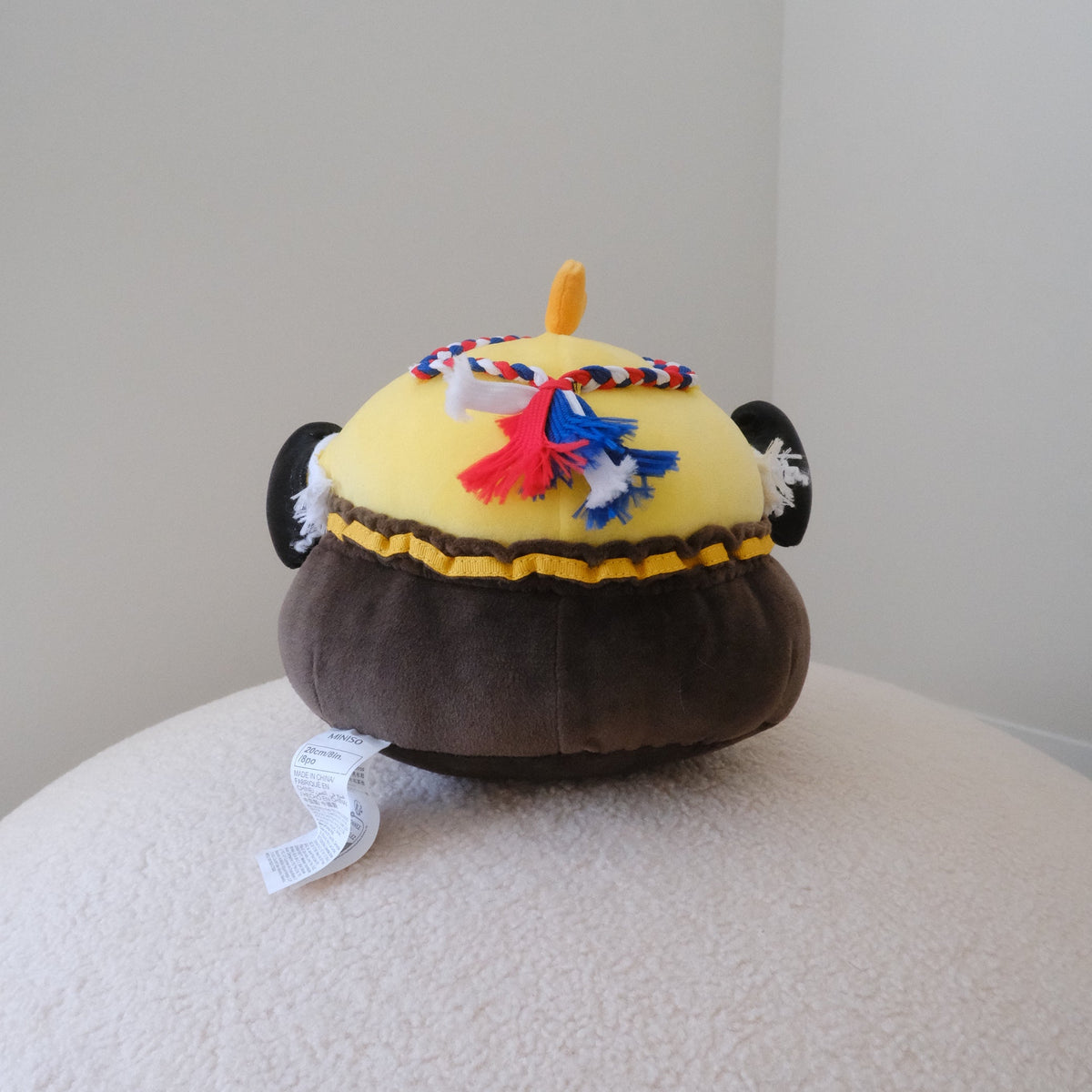A plush toy shaped like a chicken, featuring a dark brown shorts, a yellow body, and a red, white, and blue braided headband, displayed on a textured cream surface against a plain light gray background.