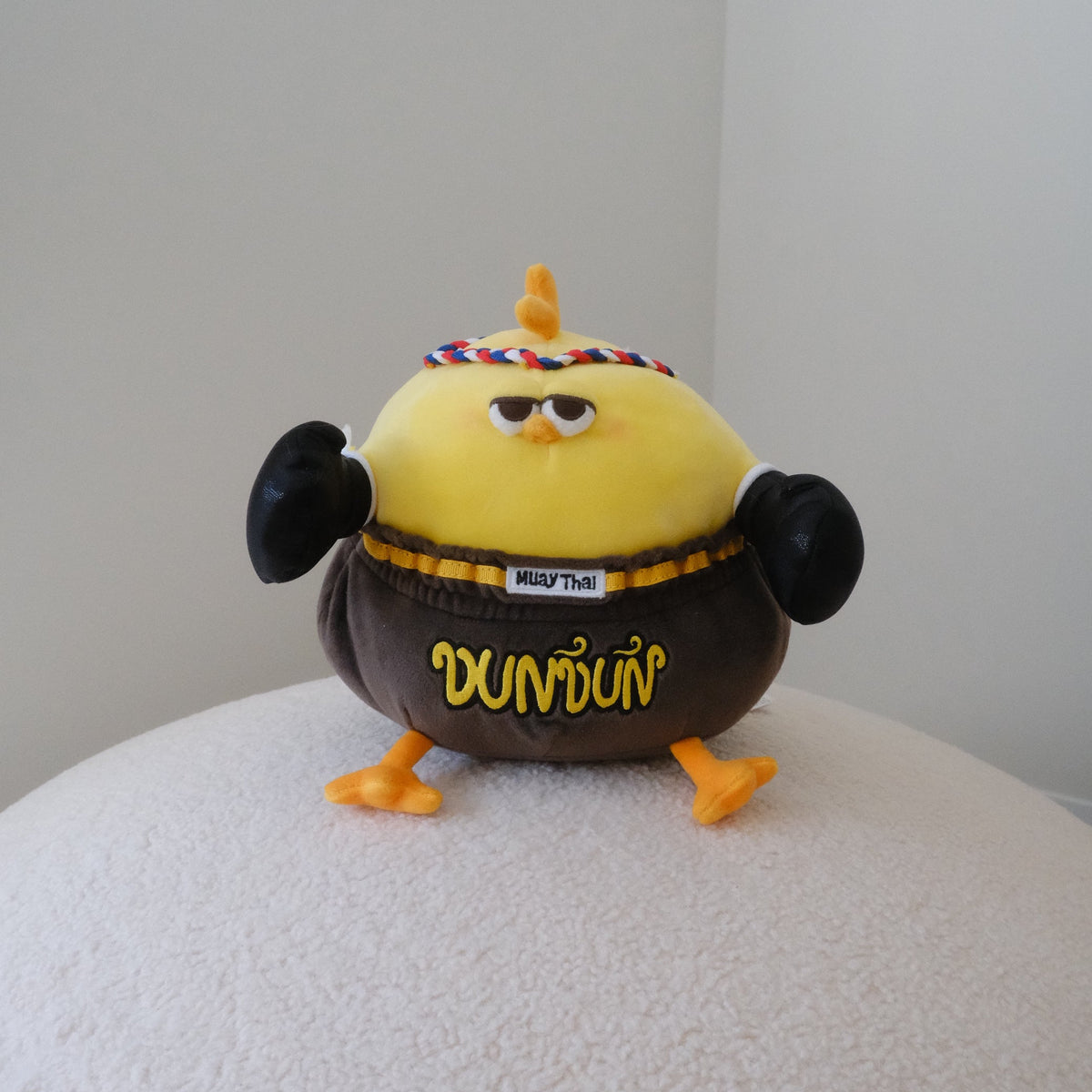 A small yellow chicken plush toy with black boxing gloves, a red and blue headband, and wearing brown shorts printed with the word 'DUNDUN' in yellow, standing on a textured white surface against a plain light gray wall.