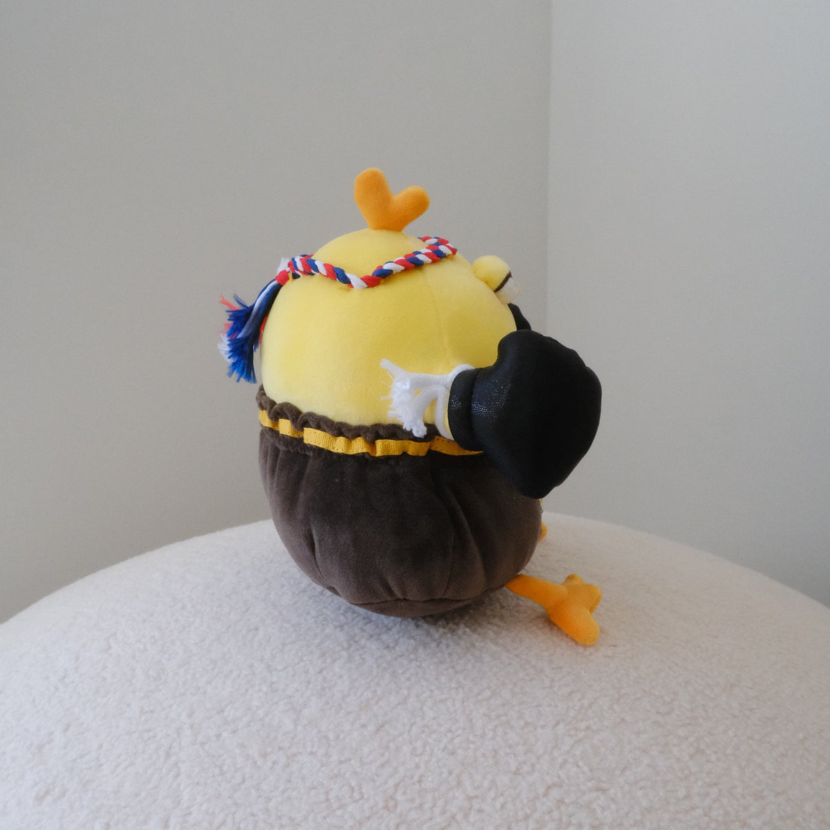 A small plush chicken toy shown from the back on a round white textured surface against a plain light gray wall. The toy has a yellow head, dark brown body, black hat, orange beak and feet, and a multicolor braided headband.
