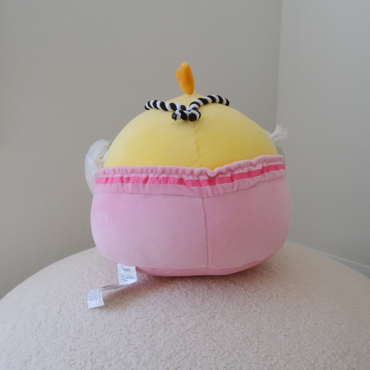 A back view of a plush chicken toy featuring a yellow body with a small orange crown and black-and-white striped headband, set in pink shorts with a ruffled trim, resting on a textured cream surface against a plain light gray background.