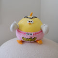 A yellow cartoon bird plush toy wearing pink shorts with 'MUAY THAI' and 'DUNDUN' text, sitting on a white textured surface against a plain light gray wall.