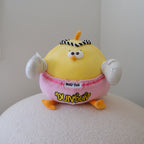 A yellow cartoon bird plush toy wearing pink shorts with 'MUAY THAI' and 'DUNDUN' text, sitting on a white textured surface against a plain light gray wall.