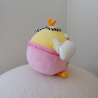 A round yellow plush toy with a pink skirt and orange feet, sitting on a textured white surface against a plain light gray wall.