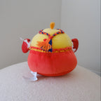 A small plush toy chicken with a yellow body, red shorts, and decorative multicolor braided detail, sitting on a textured white surface against a plain light gray wall.
