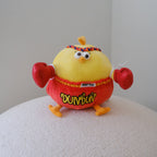 A yellow chicken plush toy wearing red boxing gloves and shorts with Thai text, sitting on a textured white surface against a plain light gray wall.