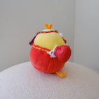 A small plush toy shaped like a yellow chicken wearing a red muay thai outfit and colorful headband, sitting on a textured white surface against a plain light gray wall.
