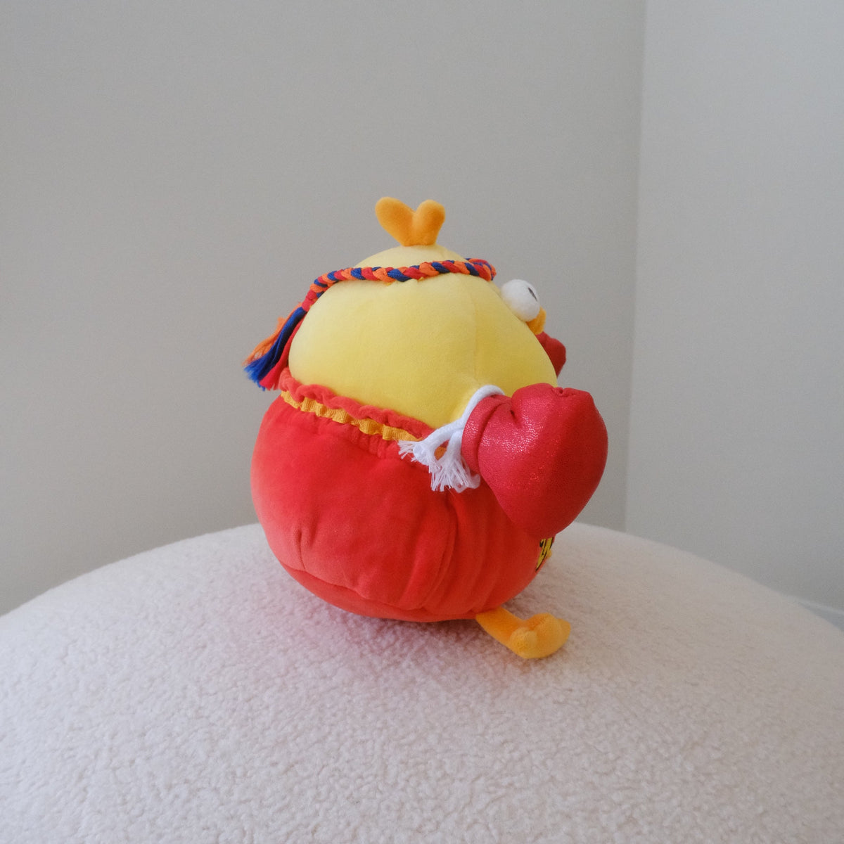 A small plush toy shaped like a yellow chicken wearing a red muay thai outfit and colorful headband, sitting on a textured white surface against a plain light gray wall.