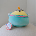 A plush toy shaped like a chicken with a yellow body, turquoise shorts with a pink stripe, black-and-white striped band, and small orange beak detail, photographed indoors on a light surface against a plain wall.