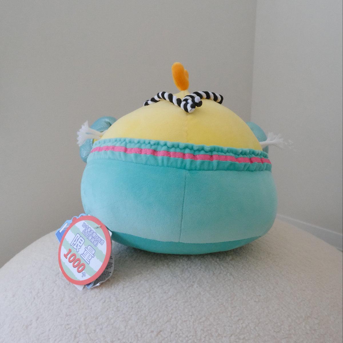 A plush toy shaped like a chicken with a yellow body, turquoise shorts with a pink stripe, black-and-white striped band, and small orange beak detail, photographed indoors on a light surface against a plain wall.