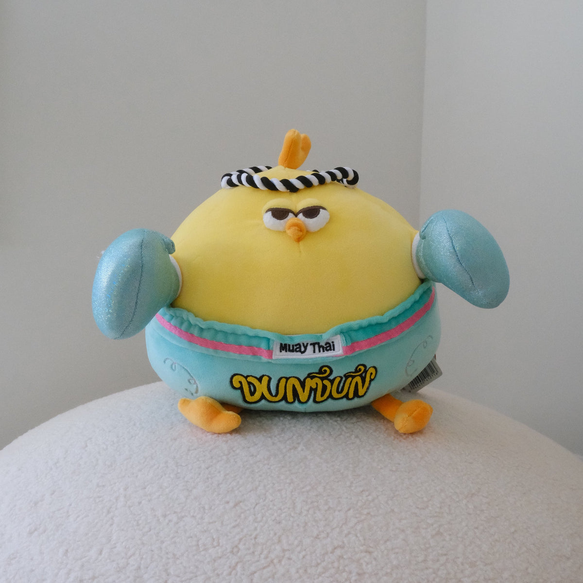 A yellow plush toy chicken with turquoise boxing gloves, wearing a black-and-white striped headband and a light turquoise shorts with the word 'DUNDUN' in yellow, sitting on a white textured surface against a plain light gray wall.