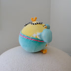 A round, plush toy chicken with a yellow head, light turquoise shorts, orange feet and beak, and black-and-white striped hat, sitting on a textured white surface against a plain light gray wall.