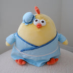 A yellow plush chicken toy wearing a light blue robe and matching blue hat, sitting on a white textured surface against a plain light gray wall.