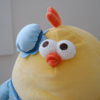 A close-up of a yellow plush toy with a round body, white embroidered eyes, an orange beak, a small orange crest on top, and a light blue bonnet with a blue band.
