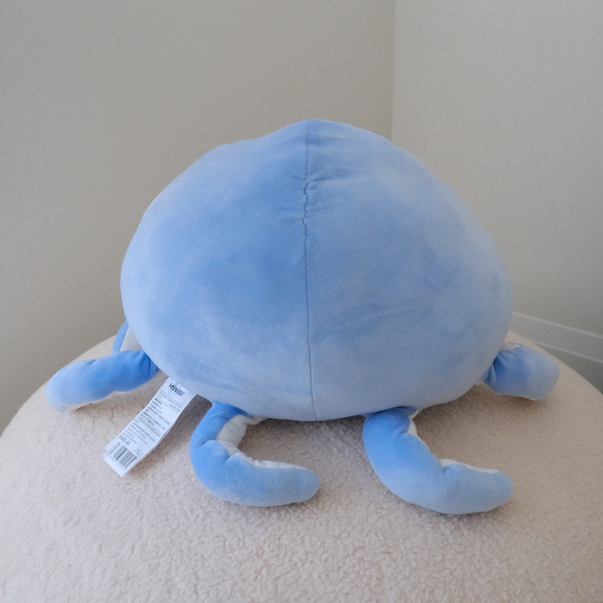 A light blue octopus plush toy with a white product tag still attached, resting on a textured beige surface against a plain beige wall background.