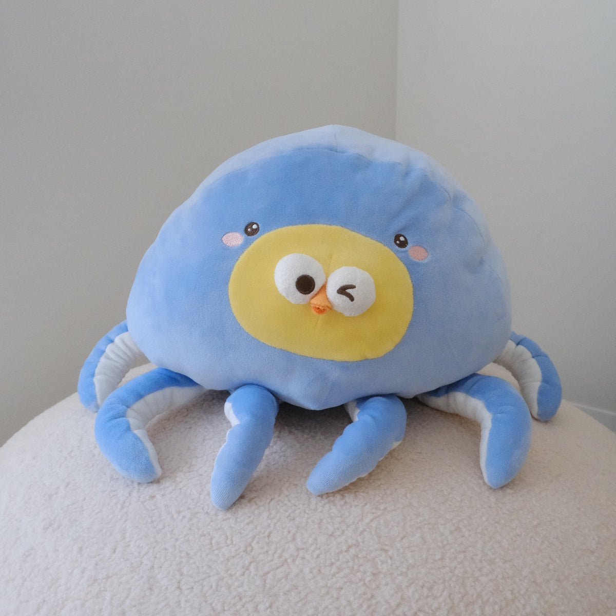 A cute blue octopus plush toy with a yellow face, resting on a textured white surface against a plain light gray wall.