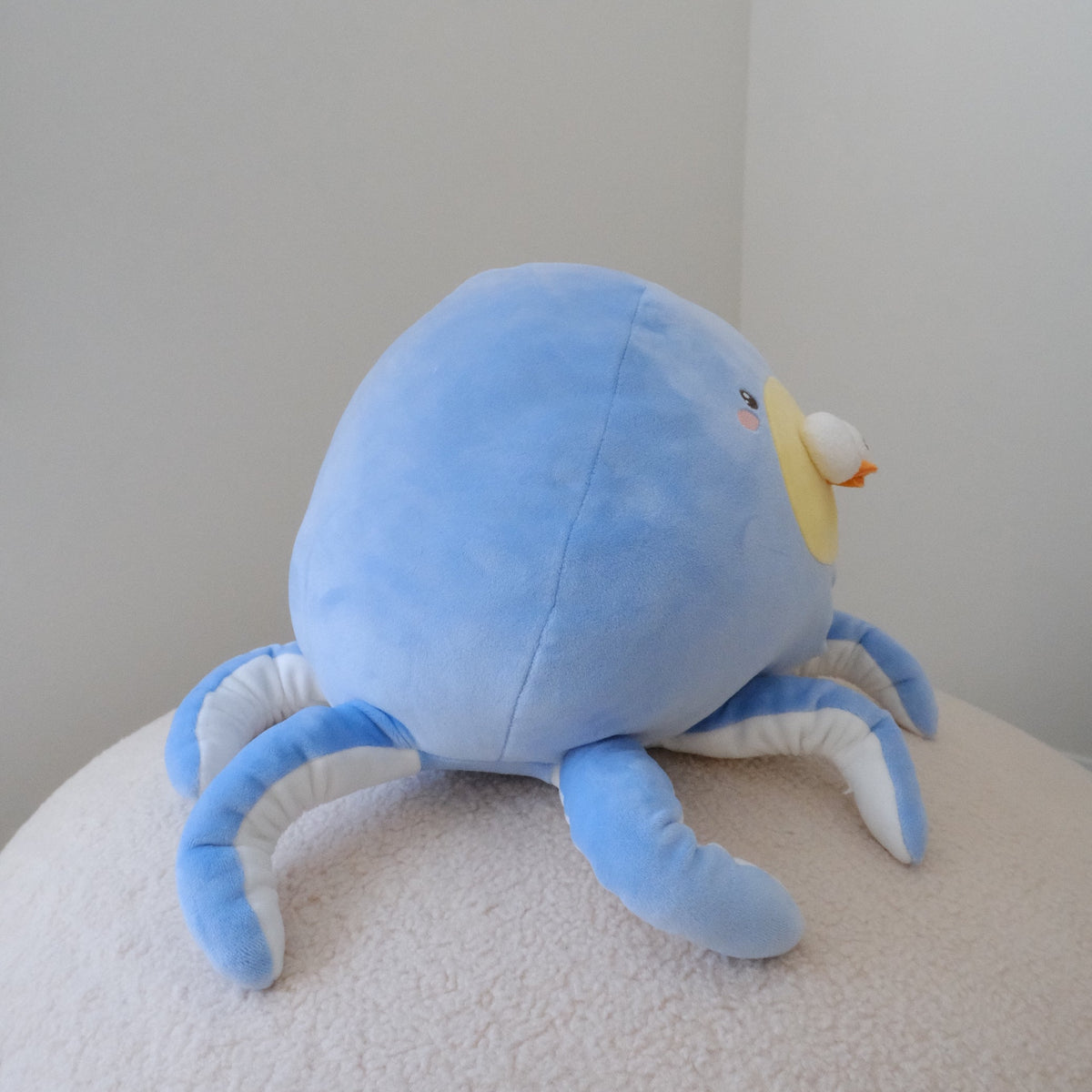 A light blue octopus plush toy with white and blue striped tentacles and a small yellow beak, sitting on a textured cream-colored surface against a plain light gray background.