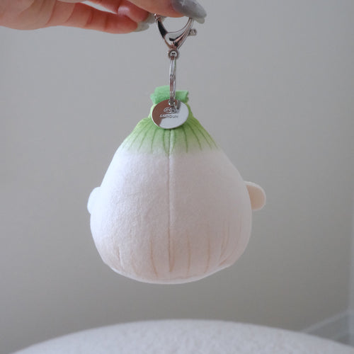 A hand holds a small plush character keychain shaped like a cute white and green onion with orange feet and beak, attached to a silver metal clasp.