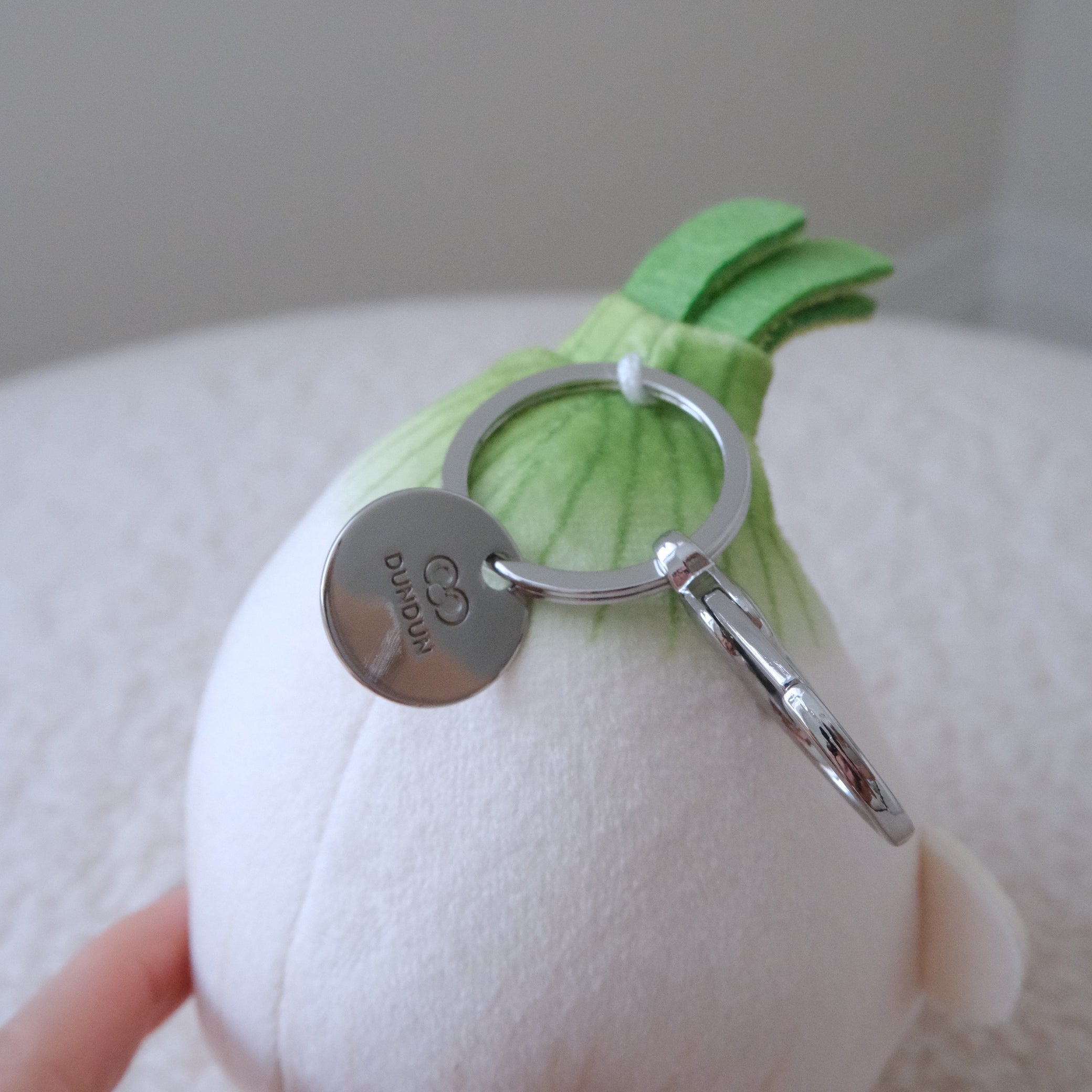 A white plush onion keychain with green fabric leaves and a silver metal keyring and key tag, held by a hand against a soft neutral background.
