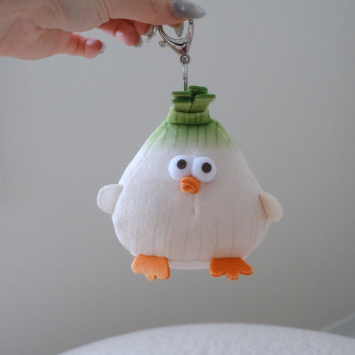 A hand holds a small plush character keychain shaped like a cute white and green onion with orange feet and beak, attached to a silver metal clasp.