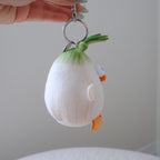 A hand holds a small plush character keychain shaped like a cute white and green onion with orange feet and beak, attached to a silver metal clasp.